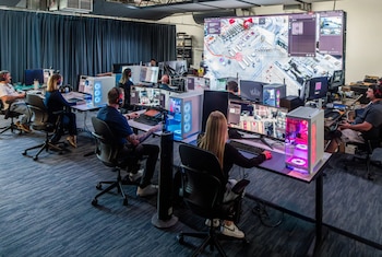 Varias personas con auriculares monitorean pantallas de computadora en un centro de control con una gran pantalla mural que muestra una vista aérea