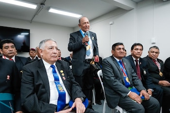 Hombre en traje con micrófono habla ante varios hombres sentados en un salón. Llevan trajes, corbatas, medallas y bandas. Se ven cajas rotuladas "SENADOR"