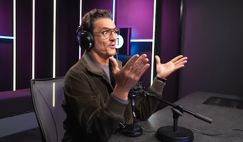 Hombre de mediana edad con gafas y auriculares habla en un micrófono en un estudio de radio, gesticulando con las manos y con luces de neón de fondo