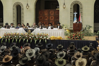 Un panel de personas sentadas en una mesa con carteles de nombres y arreglos florales. Un orador en un podio con banderas. Una audiencia con sombreros