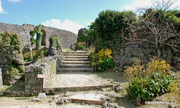Paredes de piedra, arco de piedra, escaleras de piedra, vegetación verde, árboles con flores rosadas, flores amarillas, cartel con texto en japonés, cielo azul