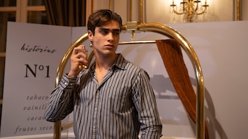 Un joven de pelo oscuro y camisa de rayas se rocía perfume. Detrás hay un cartel blanco con "historias N°1" y un carrito dorado de equipaje