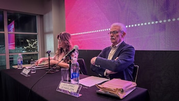 Un hombre con bigote y gafas, Guillermo Schavelzon, sentado con los brazos cruzados junto a una mujer, Verónica Abdala, en una mesa con micrófonos y botellas de agua, frente a una pantalla LED morada