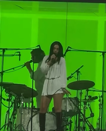 Una mujer con cabello oscuro y vestido blanco de manga larga canta en un micrófono en un escenario iluminado con luces verdes, con una batería visible en el fondo