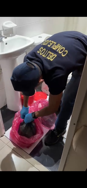 Hombre con gorra, guantes azules y camiseta "DELITOS COMPLEJOS" manipula una bolsa rosa con un objeto oscuro, junto a un fregadero y un inodoro