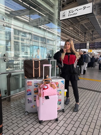 Wanda Nara, con chaqueta negra y roja, junto a tres maletas (Louis Vuitton, Rimowa, rosa) en una estación moderna con paredes de cristal y un cartel de "Elevator"