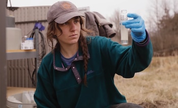 Una mujer con gorra y guantes azules sostiene un vial transparente con un líquido blanco, examinándolo atentamente al aire libre