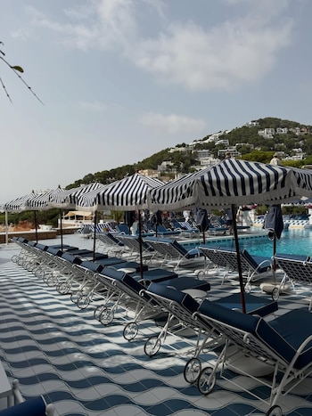 Una fila de sombrillas a rayas azules y blancas, tumbonas azules con estructura blanca, piscina, suelo con patrón ondulado azul y blanco, colina con edificios