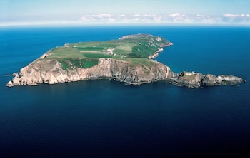 Vista superior de una isla alargada con acantilados rocosos y prados verdes, rodeada por un mar azul