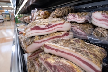 Piezas gruesas de guanciale empacado al vacío acumuladas en estantes refrigerados con un pasillo al fondo.
