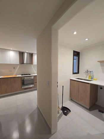 Interior de un apartamento moderno que muestra una cocina con gabinetes de madera clara y electrodomésticos, y un lavadero con fregadero y ventana