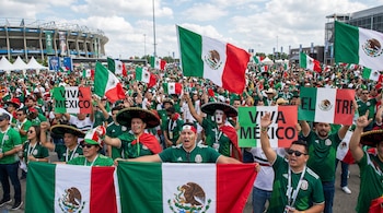 Una gran multitud de aficionados mexicanos en camisetas verdes, con sombreros y pintura facial, ondea banderas de México frente a un estadio.