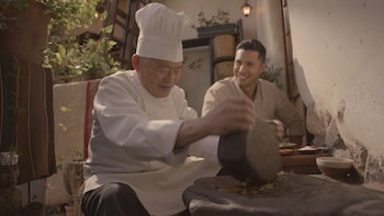 Un cocinero con gorro blanco muele ingredientes sobre una piedra mientras un hombre lo observa sonriendo en un patio con plantas