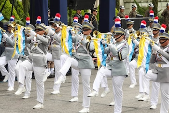 Músicos vestidos con uniformes grises, pantalones blancos y gorras con plumeros marchan y tocan trompetas con estandartes