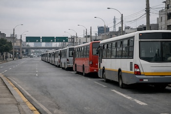 Una larga fila de buses urbanos, predominantemente blancos con uno rojo, se encuentra detenida en una avenida de Lima bajo un cielo gris nublado, sin personas visibles.
