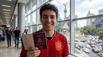 Joven sonriendo, con camiseta de España, muestra pasaporte en aeropuerto de CDMX. Detrás, a través de la ventana, se ve el Ángel de la Independencia y la ciudad.