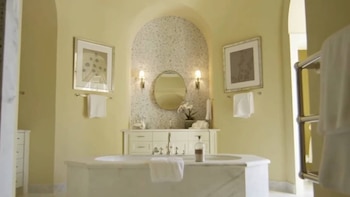Un baño elegante con bañera de mármol en el centro, un lavabo con grifos plateados bajo un espejo redondo y paredes de color crema con azulejos de mosaico