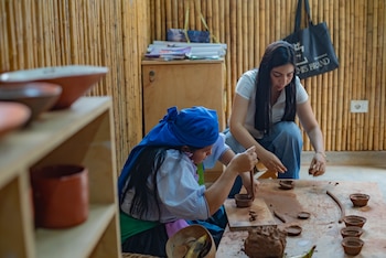 Dos mujeres sentadas en el suelo elaboran piezas de arcilla sobre una mesa de madera, con una pared de bambú al fondo y estantes con objetos de cerámica