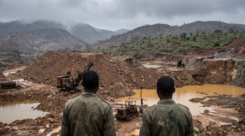 Dos hombres de espaldas con uniformes militares, mina a cielo abierto con grandes montículos de tierra, maquinaria minera antigua, charcos de agua ocre y colinas con niebla.
