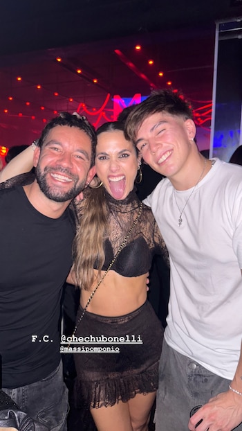 Chechu Bonelli sonríe con la lengua afuera, vestida con top y falda negros de encaje, entre dos hombres en un evento nocturno con luces rojas de fondo
