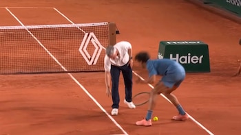 Un jugador de tenis con camiseta azul y raqueta en mano agachado en una cancha de arcilla. Un oficial con camisa blanca se inclina cerca de la red