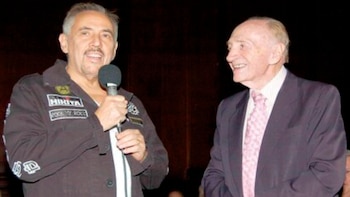 Dos hombres; uno con chaqueta oscura con parches, sostiene un micrófono. El otro viste un traje oscuro con corbata rosa y sonríe