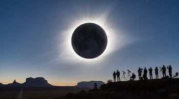 Vista panorámica de un eclipse solar total mostrando la corona brillante y prominencias rosas. Siluetas de personas observan con telescopios en un paisaje desértico.