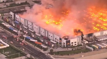 Vista aérea de un gran almacén envuelto en llamas, con espeso humo y fuego intenso en el techo. Camiones de bomberos y vehículos de emergencia actúan en la calle