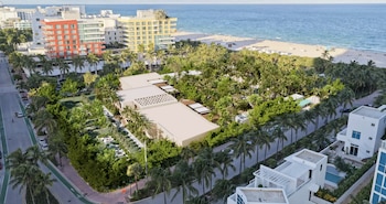Vista aérea de una zona costera con edificios modernos, calles con tráfico, abundantes palmeras y vegetación, y una playa con el océano al fondo
