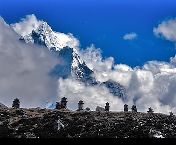 Monte Everest nevado y parcialmente cubierto por nubes blancas bajo un cielo azul brillante. En primer plano, una colina oscura con varias estructuras de piedra