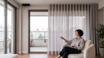 Mujer sentada en un sillón blanco sosteniendo un control remoto frente a una gran ventana con cortinas grises y vista a edificios.