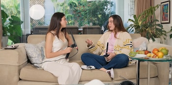 Dos mujeres conversan sentadas en un sofá beige con micrófonos en mano, junto a una mesa de vidrio con frutas y plantas al fondo