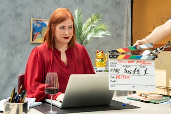 Mujer pelirroja con blusa roja sentada en un escritorio, trabajando en una laptop plateada, con una claqueta blanca de producción de Netflix frente a ella