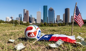 Una pelota del Mundial 2026, bandera de EE. UU., bandera de Texas y billetes de dólar en la hierba, con el horizonte de Houston de fondo bajo un cielo azul claro