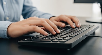 Primer plano de las manos de una persona escribiendo en un teclado de computadora negro. La persona viste una camisa azul clara.