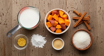 Vista superior de una mesa de madera con camote cocido en cubos, leche de coco en jarra, canela en rama, coco rallado, fécula, y dos pequeños cuencos.