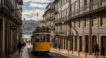 Tranvía amarillo en calle adoquinada con edificios tradicionales, personas caminando, postes de luz, cables aéreos, un río y un puente a lo lejos, cielo nublado.