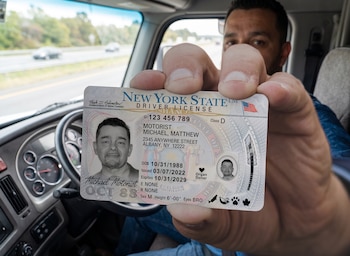 Un hombre en el asiento del conductor de un vehículo sostiene una licencia de conducir del estado de Nueva York. Una carretera borrosa se ve al fondo