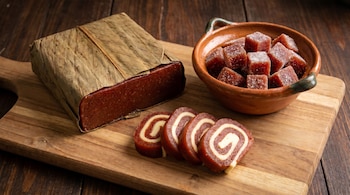 Una tabla de madera muestra una barra de dulce de guayaba envuelta en hojas, cubos de guayaba azucarados en un cuenco de barro y varias rebanadas con queso en espiral.