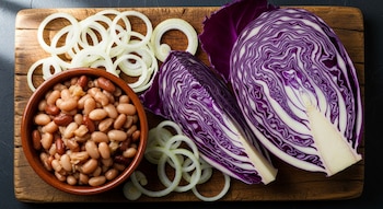 Vista cenital de porotos cocidos en un tazón de cerámica, dos mitades de repollo morado fresco y aros de cebolla cortada en una tabla de madera.