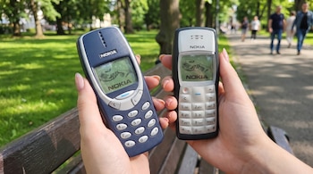 Manos sosteniendo dos teléfonos móviles Nokia, modelos 3310 azul y 1100 plateado, con pantallas mostrando un juego. Fondo de parque con césped y árboles.
