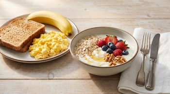 Bodegón de desayuno con un bol de yogur, avena, frutos rojos, miel y nueces; un plato con huevos revueltos, tostadas y plátano; y cubiertos sobre servilleta.