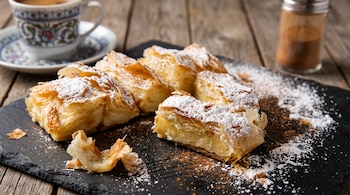 Trozos de bougatsa espolvoreados con azúcar y canela sobre una tabla de piedra negra, junto a una taza de café y una mesa de madera.