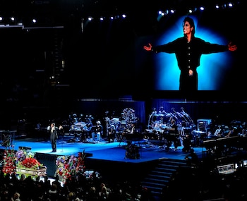 Un escenario oscuro con una pantalla gigante que muestra una imagen de Michael Jackson, un artista cantando, una banda y un féretro dorado con flores