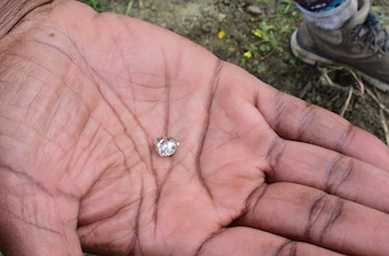 Primer plano de una palma de mano sucia sosteniendo un diamante brillante y claro. Al fondo, se ve un suelo terroso y parte de un zapato