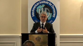 Hombre en traje y gafas, parado frente a un podio con placa National Doral Miami. Detrás, un logo azul del InterAmerican Institute for Democracy
