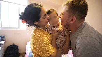Una mujer besa la mejilla de una niña mientras un hombre mira a la niña. La niña está en brazos de la mujer. Los tres están en una habitación