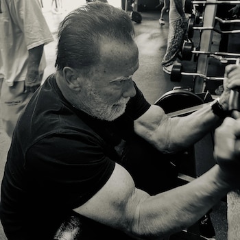 Imagen en blanco y negro de Arnold Schwarzenegger, un hombre mayor con barba y camiseta negra, realizando un ejercicio de bíceps en el gimnasio