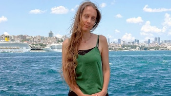 Retrato de Shelly Renee Kittleson, mujer con cabello castaño largo y ondulado, camiseta de tirantes verde, frente a un mar y una ciudad con edificios