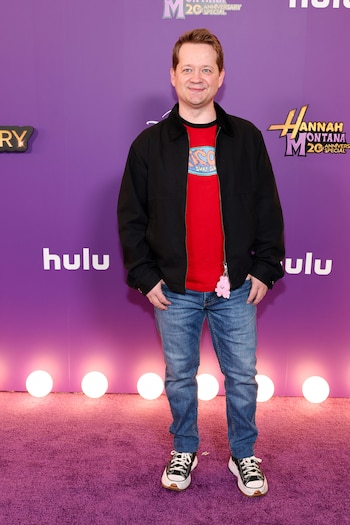 Jason Earles, un hombre de piel clara y cabello castaño rojizo, sonríe en un evento. Viste chaqueta negra, camiseta roja, jeans y zapatillas deportivas. Fondo morado con logos